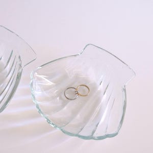 May include: Two clear, seashell-shaped glass dishes. One dish contains two rings, one gold and one silver. The dishes are on a white surface, with soft lighting creating shadows and highlights.