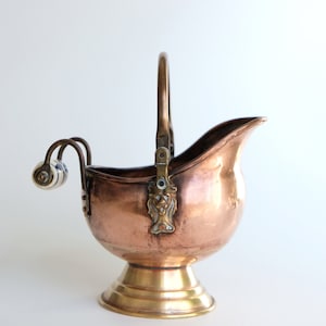 Könnte beinhalten: Ein Eiseimer aus Kupfer und Messing mit einem Griff und zwei Porzellanknöpfen. Der Eimer hat ein dekoratives Design an der Seite.