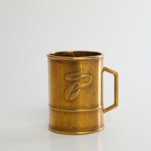 Könnte beinhalten: Eine kleine, goldfarbene Metalltasse mit einem Kaffeekorn-Design. Die Tasse hat einen Henkel und einen abgerundeten Boden.
