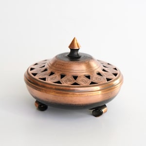 Puede incluir: Un quemador de incienso de cobre con un diseño decorativo y una tapa puntiaguda. El quemador tiene tres patas y está sobre una superficie blanca.