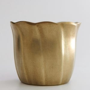 Könnte beinhalten: Ein goldfarbener, geriffelter Pflanztopf mit gewelltem Rand. Die metallische Oberfläche verleiht dem Keramiktopf einen Vintage-Look. Der Behälter ist für den Innen- und Außenbereich geeignet und kann eine Vielzahl von Pflanzen aufnehmen.