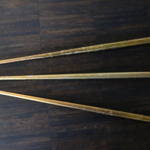 Op de afbeelding: Drie goudkleurige metalen staven met afgeronde, kogelvormige uiteinden. De staven liggen diagonaal op een donkere houtnerf oppervlak. De staven lijken vintage of antiek te zijn, met wat slijtage zichtbaar.