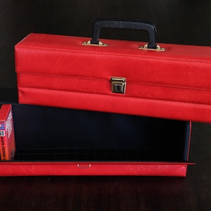 May include: A red vinyl case with a black handle and gold-coloured hardware. The case is open, revealing a black interior and a tray with several game cartridges. The case is designed to hold games or other small items.