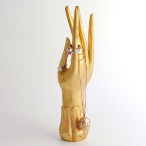 Könnte beinhalten: Ein goldfarbener, handförmiger Schmuckständer mit Ringen und einer Halskette mit einem runden Anhänger.