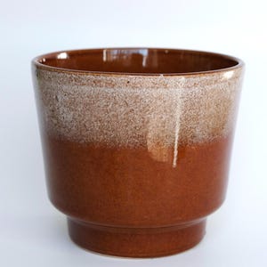 Op de afbeelding: Een bruine keramische plantenpot met een tweekleurig ontwerp. De bovenste helft van de pot heeft een gespikkelde, lichtbruine afwerking, terwijl de onderste helft effen, donkerder bruin is. De pot heeft een brede opening en een voet.