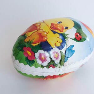 May include: A colorful Easter egg-shaped chocolate candy with a cartoon duckling design. The duckling is yellow with orange feet and a blue background. The egg is surrounded by flowers and ladybugs.