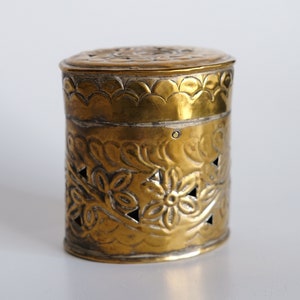 Op de afbeelding: Een gouden metalen container met een sierlijk bloemenpatroon. De container heeft een deksel en is versierd met een herhalend patroon van bloemen en bladeren.