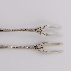 May include: Two silver forks with ornate handles. The forks are about 5 inches long and have a floral design. The forks are marked with the number "900" on the back.