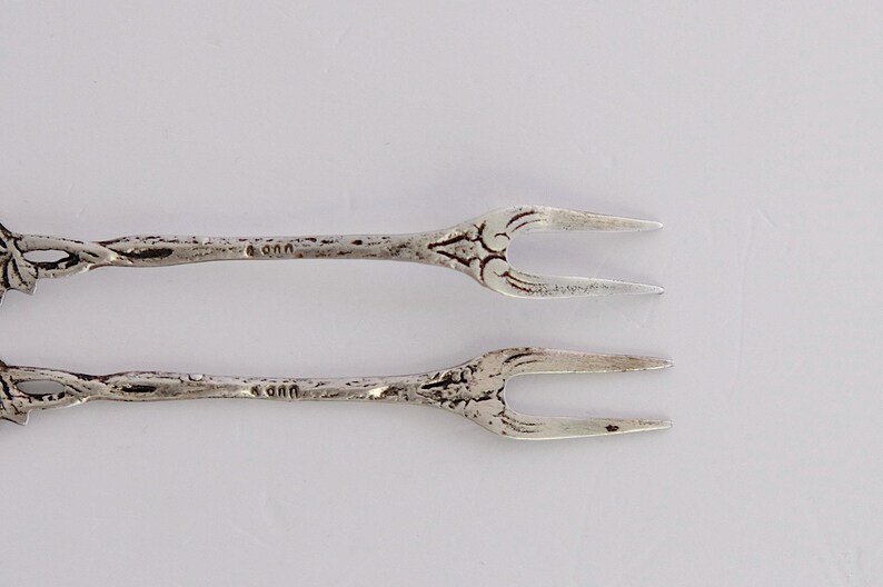 May include: Two silver forks with ornate handles. The forks are about 5 inches long and have a floral design. The forks are marked with the number "900" on the back.