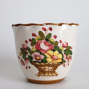 Puede incluir: Un macetero de cerámica blanca con un borde marrón y un diseño floral. El diseño presenta flores rosas, amarillas y verdes en una cesta marrón.