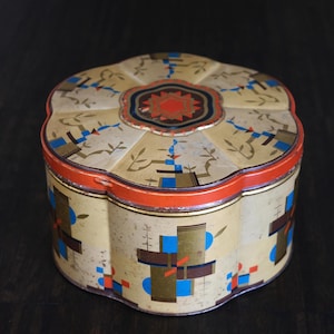 Könnte beinhalten: Vintage, blumenförmige Blechdose mit cremefarbenem Hintergrund. Die Dose hat ein geometrisches Design in Rot, Blau und Gold. Der Deckel hat ein zentrales Blumendesign in Rot, Schwarz und Gold. Die Dose dient wahrscheinlich zur Aufbewahrung.