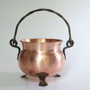 May include: A copper cauldron with a hammered texture, featuring a dark metal handle and three small feet. The handle is arched and attached to the cauldron with decorative metalwork. The cauldron has a shiny, reflective surface.