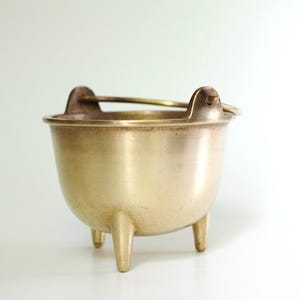 Könnte beinhalten: Ein kleiner, antiker Messingkessel mit drei kurzen Beinen und einem Griff. Der Kessel hat einen abgerundeten Körper und einen leicht ausgestellten Rand. Der Griff ist an zwei kleinen, dekorativen Halterungen auf beiden Seiten des Rands befestigt. Das Messing hat einen warmen, goldenen Farbton.