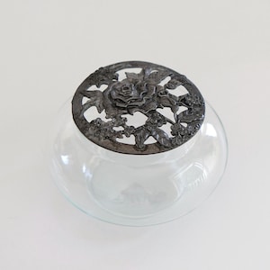 May include: A clear glass vase with a silver metal lid featuring a floral design with a rose in the center.