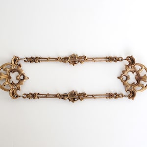 Könnte beinhalten: Zwei verzierte goldene Dekorationsstücke aus Metall mit komplizierten Designs. Die Stücke sind durch eine Kette verbunden und können zum Aufhängen eines Bildes oder Spiegels verwendet werden.