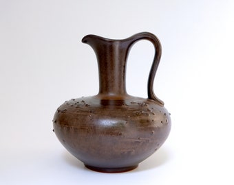 May include: A brown ceramic pitcher with a textured surface and a rounded body. The pitcher has a narrow neck and a curved handle.