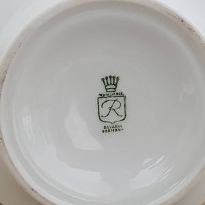 May include: Close-up of a white porcelain plate with a green logo. The logo features a crown, a shield with the letter "R" inside, and the text "Wunsiedel Bavaria Germany".