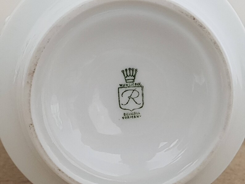 May include: Close-up of a white porcelain plate with a green logo. The logo features a crown, a shield with the letter "R" inside, and the text "Wunsiedel Bavaria Germany".
