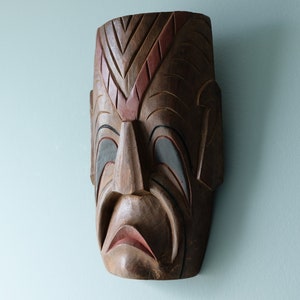 Op de afbeelding: Een houten masker met ingewikkelde houtsnijwerk en beschilderde details. Het masker toont een menselijk gezicht met een droevige uitdrukking. Het masker is bruin, rood en grijs.