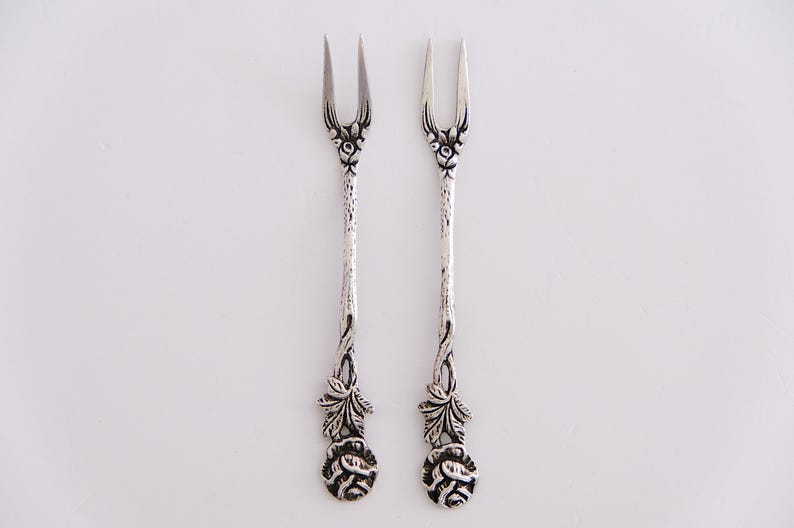 May include: A pair of silver-plated, ornate forks with floral designs. The forks have a rose design at the base of the handle and a leaf design on the stem.