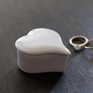 Op de afbeelding: Een witte, hartvormige ringendoos van keramiek met twee gouden ringen op een donker houten oppervlak.