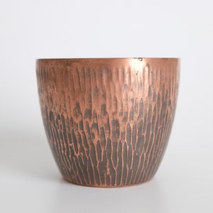 May include: A hammered copper cup with a textured surface. The cup is a light copper color at the top and darkens to a brown color at the bottom.