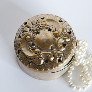 May include: A silver and gold round trinket box with an ornate design on the lid. A strand of white pearls is next to the box.