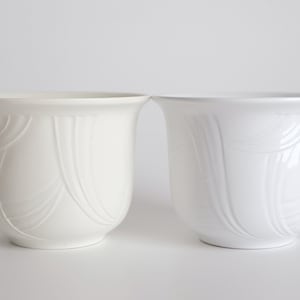 Könnte beinhalten: Zwei weiße Keramik-Blumentöpfe mit einem dekorativen Prägemuster. Die Töpfe sind in Form und Größe ähnlich.