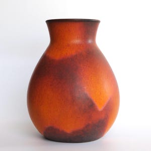 Puede incluir: Un jarrón de cerámica con cuerpo redondeado y cuello estrecho. El jarrón presenta un degradado de colores, pasando de una base marrón oscuro a un tono naranja vibrante y marrón rojizo. El borde es de color marrón oscuro.