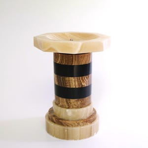 Puede incluir: Un pedestal decorativo con una parte superior octogonal de color claro. La columna presenta bandas alternas de vetas de madera, negro y piedra clara. La base es una serie de capas circulares apiladas. El diseño general es una mezcla de materiales naturales.