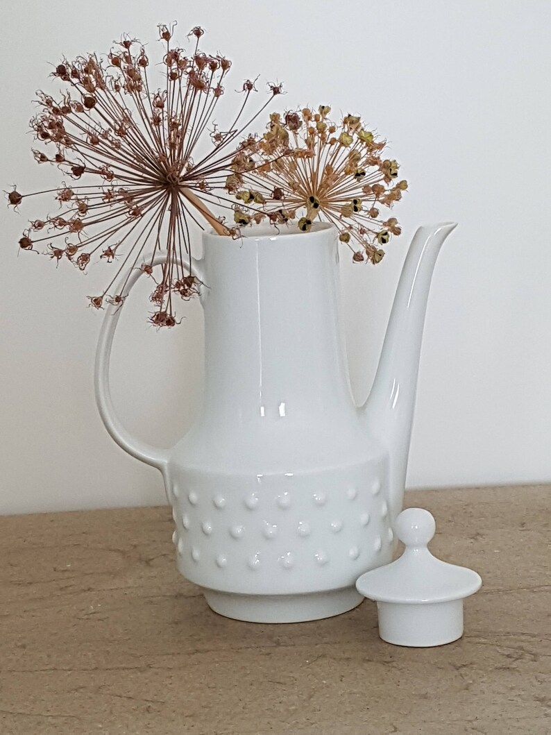 May include: White ceramic coffee pot with a textured design and a matching lid. The pot has a rounded body and a spout. The lid has a knob on top. The pot is sitting on a wooden surface.