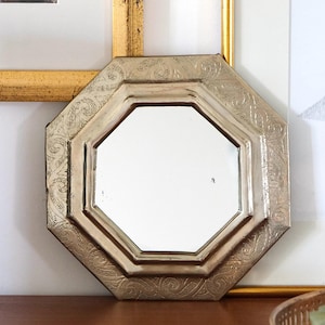 May include: Octagonal silver-toned decorative mirror with an ornate design. The mirror has a beveled edge and a clear reflection.
