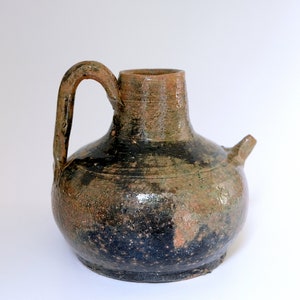 Op de afbeelding: Een bruine keramische kan met een rond lichaam en een korte, smalle hals. De kan heeft een handvat aan de ene kant en een tuit aan de andere kant. De glazuur is onregelmatig, met gebieden van donkerbruin, lichtbruin en groen.