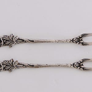 May include: Two silver-toned, ornate, rose-shaped, small forks with a decorative floral design.