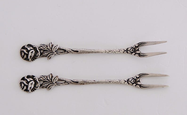 May include: Two silver-toned, ornate, rose-shaped, small forks with a decorative floral design.