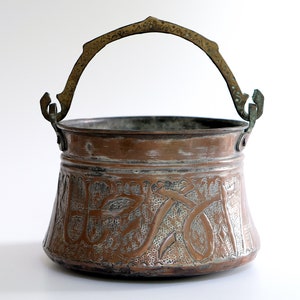 May include: An antique copper pot with an ornate engraved design and a brass handle. The pot has a wide, round base and a slightly flared rim.