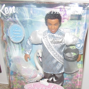 Puede incluir: Muñeco Ken vestido como el Príncipe Daniel de Barbie del Lago de los Cisnes. Lleva un atuendo plateado y azul con una banda plateada y una corona. Sostiene un cisne blanco con detalles rosas. La caja tiene el texto "Lovely Swan for you!" y "Barbie del Lago de los Cisnes".