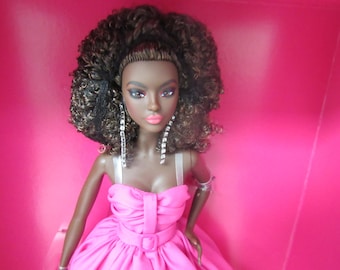 May include: A fashion doll with dark skin, wearing a bright pink dress with a belt. The doll has curly brown hair and long, dangling silver earrings. The background is a solid pink color.