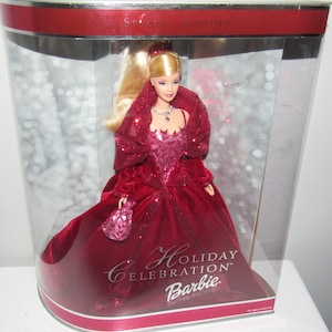 Puede incluir: Una muñeca Barbie de terciopelo rojo con un vestido de terciopelo rojo, un collar de oro y un bolso rojo. La muñeca está en una caja de plástico transparente con las palabras "Holiday Celebration Barbie" en la parte delantera.