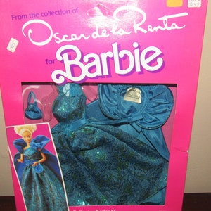 Może przedstawiać: Strój dla lalki Barbie zaprojektowany przez Oscara de la Rentę. Strój to turkusowa suknia z dopasowaną peleryną i małą torebką. Pudełko jest różowe i zawiera logo Barbie oraz logo Oscara de la Renty. Tekst "Collector Series V" jest nadrukowany na spodzie pudełka.