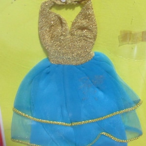 Puede incluir: Un vestido de muñeca con un corpiño estilo halter en color dorado brillante y una falda azul de varias capas. La falda consta de una base azul sólida cubierta por una capa de tul azul, ambas con un borde fino de color dorado.