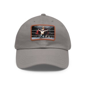 Puede incluir: Una gorra de béisbol gris con un parche marrón que muestra a un luchador con una capa roja celebrando una victoria en un ring de lucha libre.