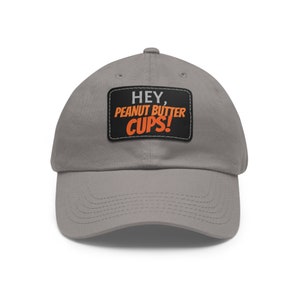 Può includere: Un cappello da baseball grigio con una toppa nera che dice "HEY, PEANUT BUTTER CUPS!" in lettere bianche e arancioni.