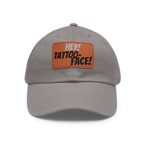 Può includere: Un cappello da baseball in cotone grigio con una toppa arancione che dice "Hey! Tattoo-Face!" in testo bianco e nero.