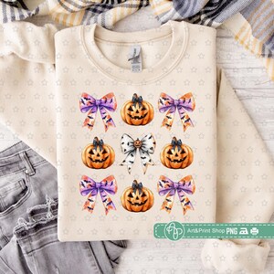 May include: A beige sweatshirt with a repeating pattern of orange and white jack-o'-lantern faces and purple and orange bows. The bows have a watercolor effect.