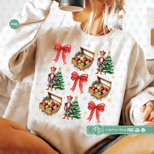 Puede incluir: Sudadera blanca con un patrón repetido de lazos rojos, cascanueces, árboles de Navidad y cajas llenas de adornos coloridos. El patrón está sobre un fondo blanco.