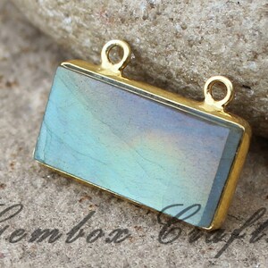 May include: A gold-toned rectangular pendant with a light blue iridescent gemstone. The pendant has two jump rings for attaching to a chain or cord.