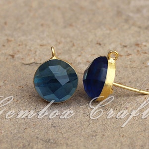 May include: Two gold-toned earring findings with blue glass cabochons. The cabochons are round and have a faceted design. The findings have a loop at the top and a post at the bottom.