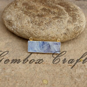 May include: A rectangular blue and white gemstone pendant with gold-toned metal accents. The pendant is on a light brown textured surface.