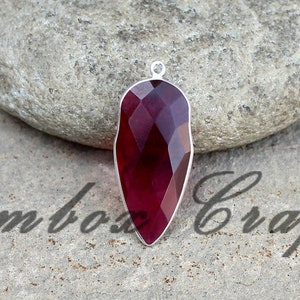 Könnte beinhalten: Ein tiefroter, facettierter Edelstein-Anhänger mit einer silbernen Öse. Der Anhänger ist tropfenförmig und vor einem grauen Hintergrund platziert.